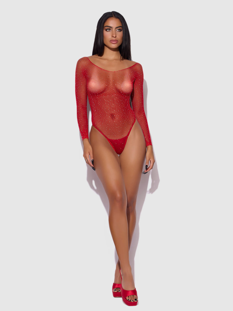 A woman stands against a plain light background, wearing the Fine Fishnet & Rhinestone Long Sleeve Teddy Bodystocking by Frederick’s of Hollywood. The sheer bodysuit is adorned with sparkling red rhinestones and features long sleeves, paired with matching red heels. She has long dark hair and a neutral expression.