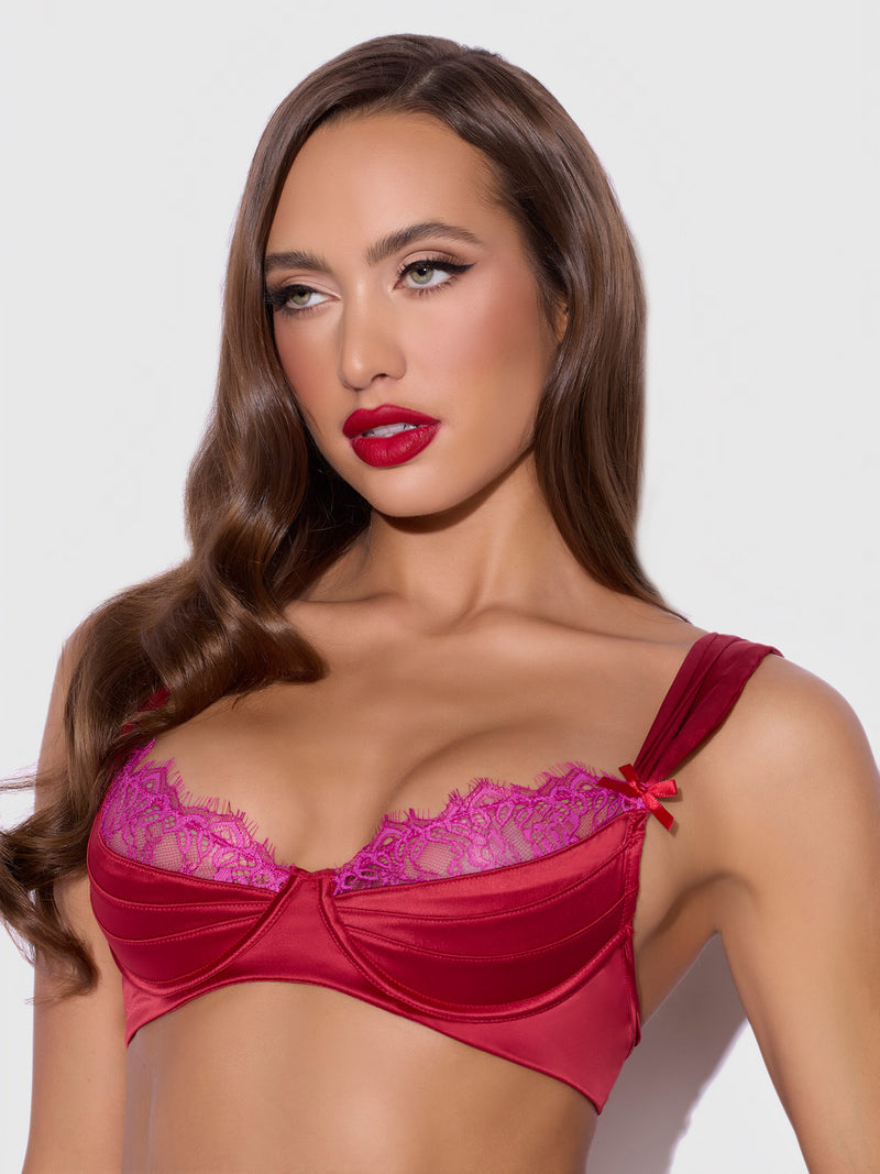 A woman with long brown hair and red lipstick models the Frederick's of Hollywood Rosalind Satin & Lace Satin Half Cup Bra, featuring red satin and purple lace trim, against a plain light background.
