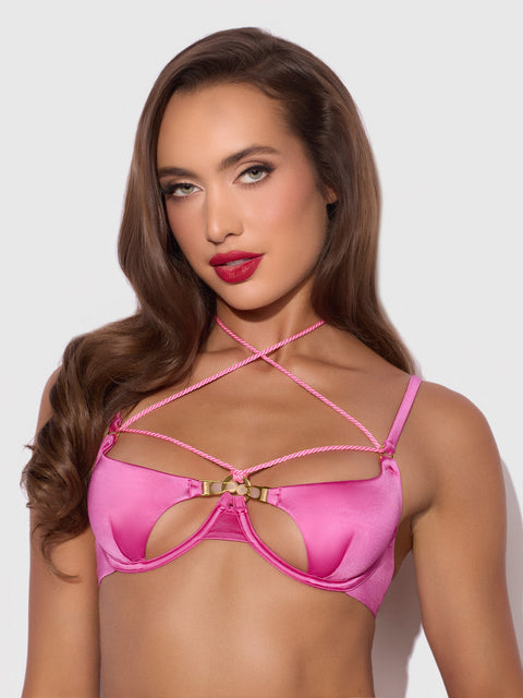 A woman with long brown hair poses in the Nadya Liquid Satin & Rope Openable Cup Bra by Frederick’s of Hollywood, featuring pink strappy details and gold accents, against a plain light background.