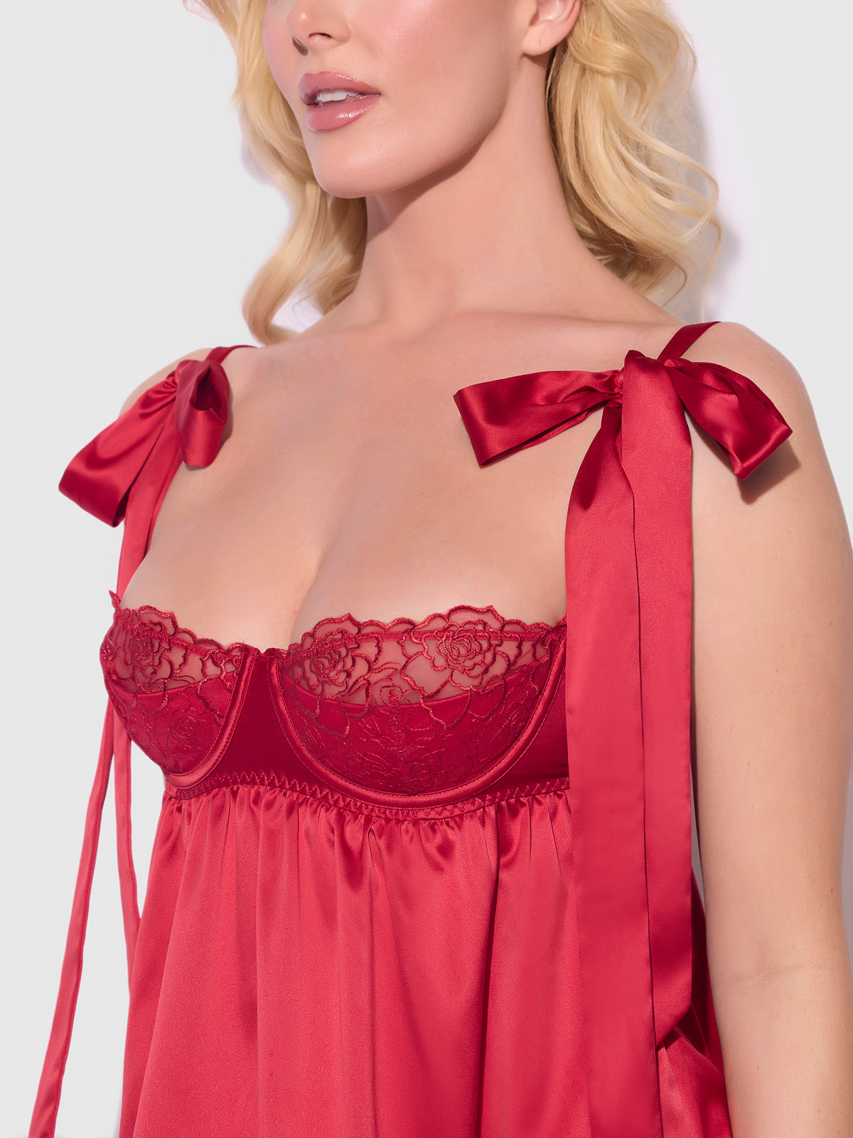 A woman with blonde hair models the Frederick's of Hollywood Melrose Satin & Embroidered Lace Underwire Babydoll Set in red, featuring lace-detailed cups and large satin bows on the straps, shown from lips to mid-torso.