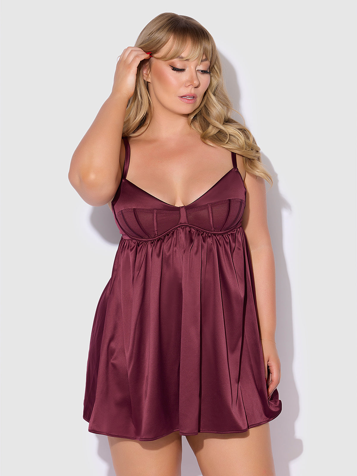 A woman models the Frederick's of Hollywood Petra Satin & Mesh Unlined Corset Cup Babydoll Set in burgundy, standing against a light gray background, touching her hair with one hand and gazing slightly downward.