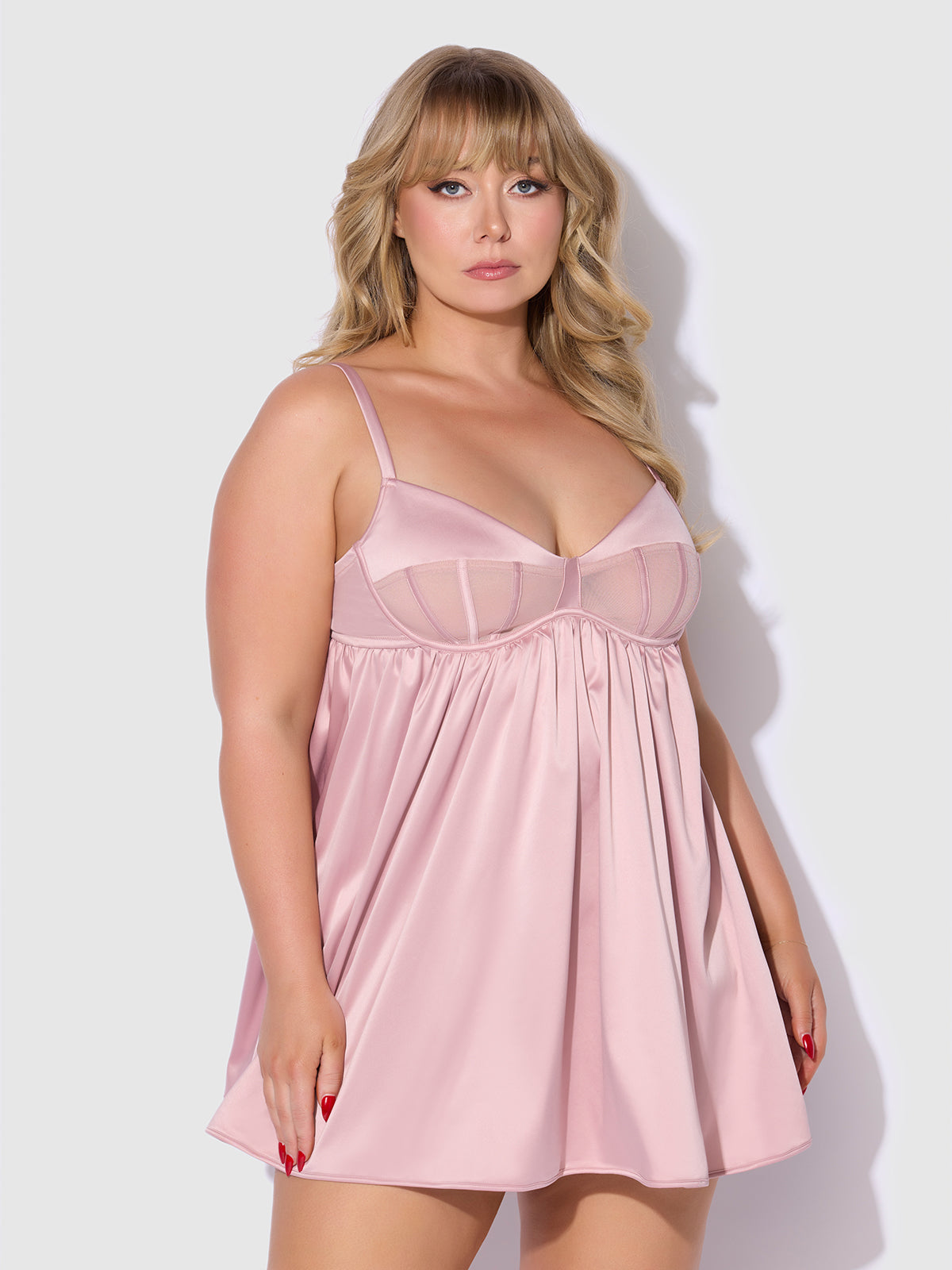A woman with long blonde hair models the Frederick's of Hollywood Petra Satin & Mesh Unlined Corset Cup Babydoll Set in light pink, featuring thin straps, standing against a plain white background.