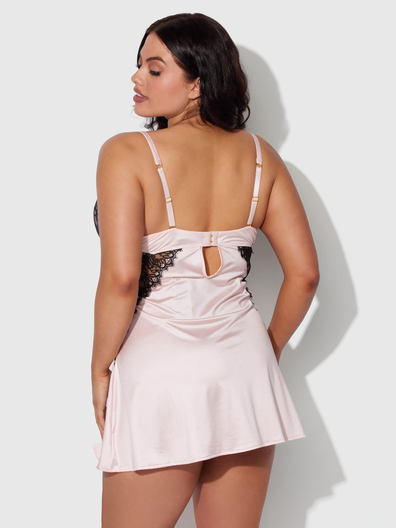 A woman poses facing away in the Frederick's of Hollywood Rising Star Plunge Push Up Babydoll in liquid satin and lace, light pink with black lace details, adjustable straps, a back cutout, and a loose mid-thigh fit.