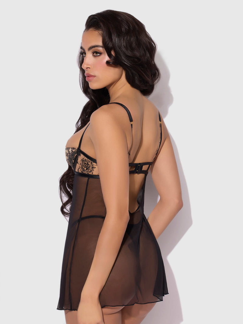 A woman with long dark hair models the Delfina Embroidered Mono Wire Babydoll by Frederick's of Hollywood, featuring lace and mesh details, posing with her back slightly turned against a plain light background.