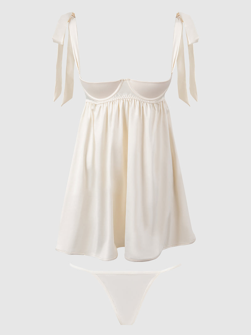 The Petra Satin Open Cup Babydoll by Frederick's of Hollywood is a cream-colored satin lingerie set with a short, flowy chemise featuring tied shoulder straps and a matching thong, shown on a plain light background for an elegant look.