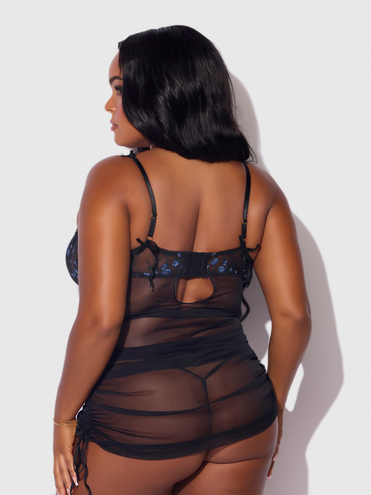 A woman with long, dark hair is seen from behind in the Frederick's of Hollywood Harlow Embroidered Stretch Mesh Chemise, featuring sheer black mesh with blue floral embroidery and adjustable straps, set against a plain light background.