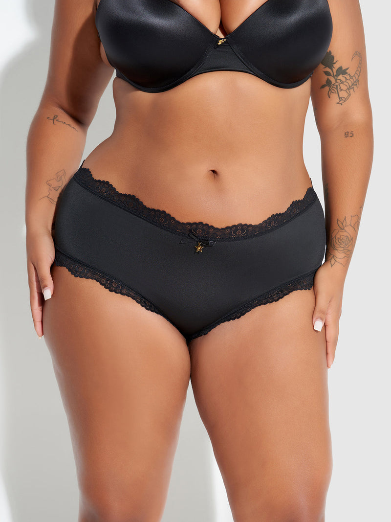 A tattooed person is shown from shoulders to thighs in Frederick's of Hollywood Jenna Micro Cheeky, featuring a black satin bra and medium-rise lace panties, standing against a neutral background.
