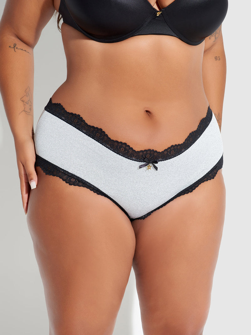 A person stands against a neutral background wearing the Frederick's of Hollywood Jenna Micro Cheeky panties in light gray with black trim and a small bow, paired with a black bra. Tattoos are visible on their arms.