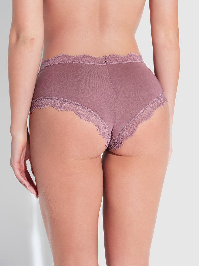 A person is seen from behind wearing Frederick's of Hollywood Jenna Micro Cheeky panties in mauve, featuring medium rise, scalloped lace edges, and a cheeky fit with moderate coverage against a softly lit plain background.