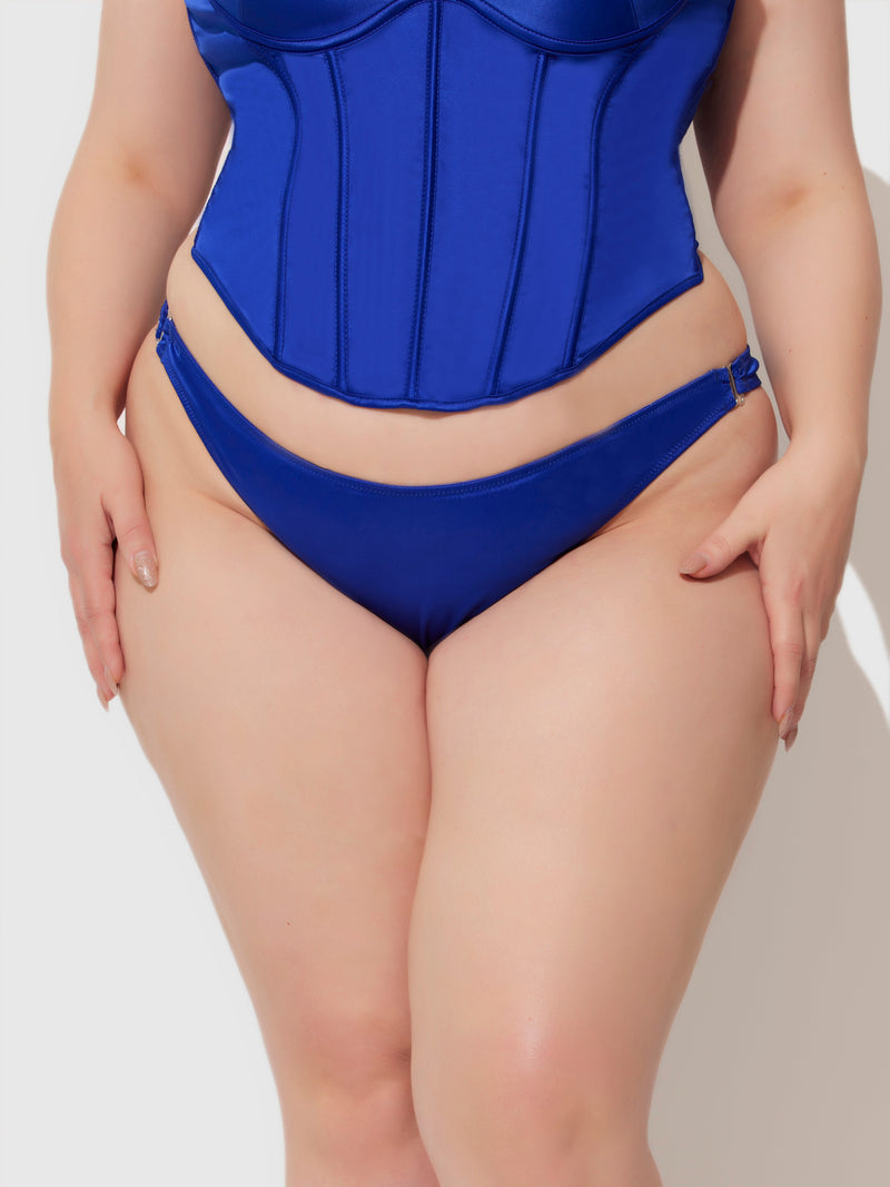 A person wears the Frederick's of Hollywood Petra Crotchless Satin Tanga, a blue silky bikini-style lingerie piece, posing with hands relaxed at their hips against a plain white background.