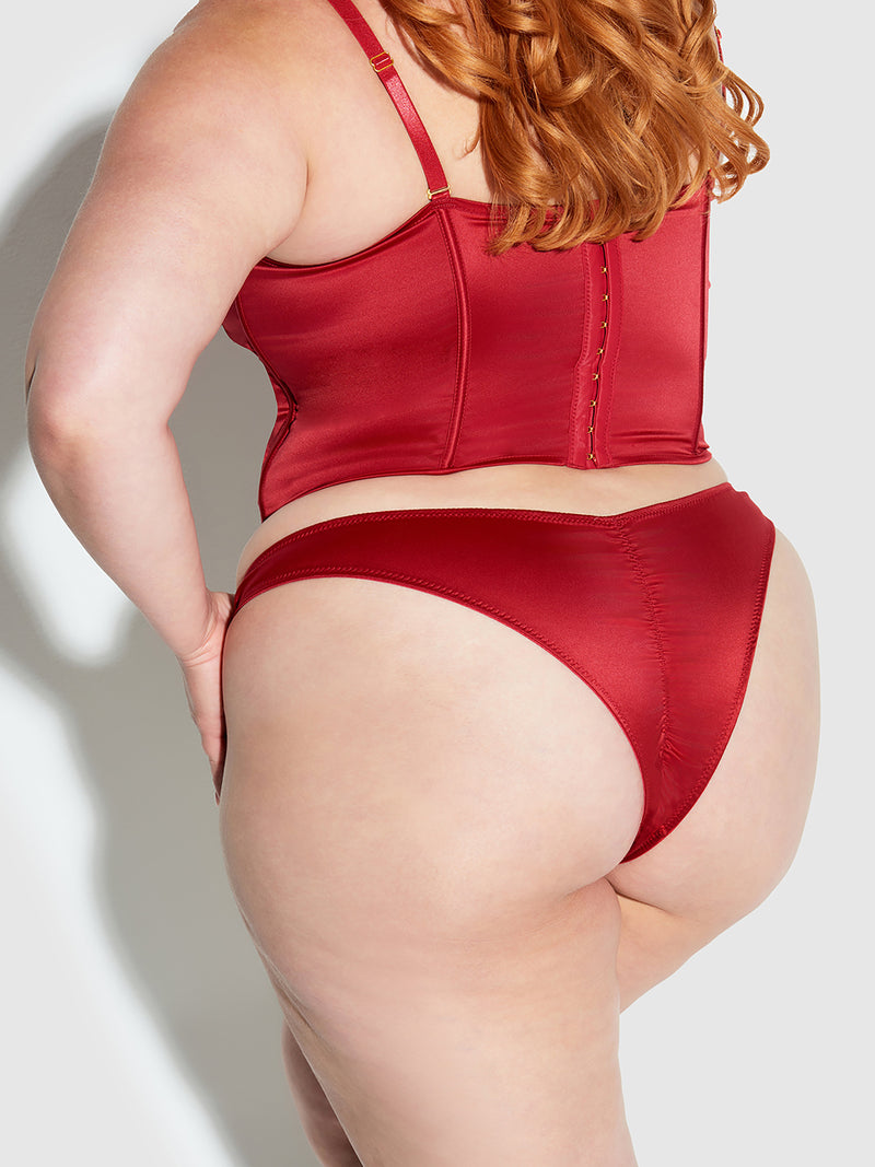 A person with long red hair poses with their back to the camera, wearing the Frederick's of Hollywood Petra Crotchless Satin Tanga. Soft lighting creates gentle shadows against a light-colored background.