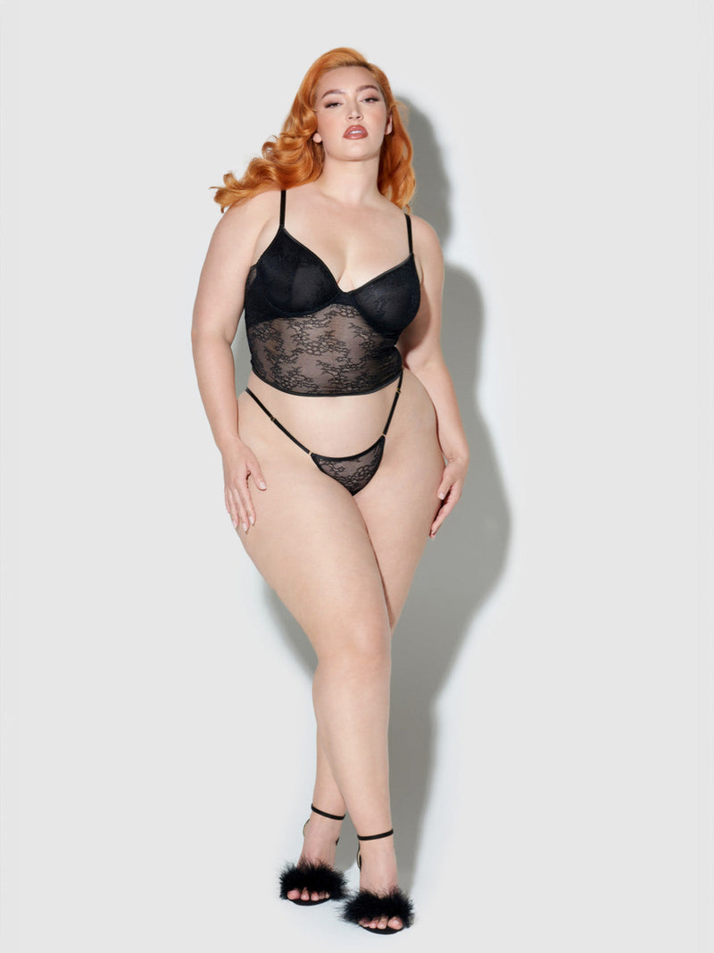 A woman with long red hair poses confidently in a Frederick's of Hollywood Chiara Crotchless Lace Thong from the 3 Pack, paired with high-heeled sandals, against a plain light gray background.