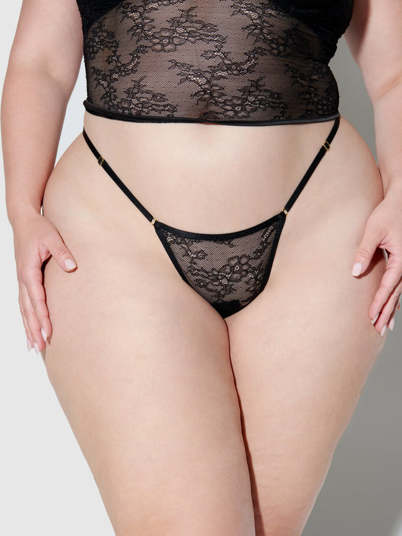 Model wears the Frederick's of Hollywood Chiara Crotchless Lace Thong 3 Pack, styled with a minimal coverage high-waisted top. The sheer black lace lingerie set is shown against a neutral background, hands resting on hips.