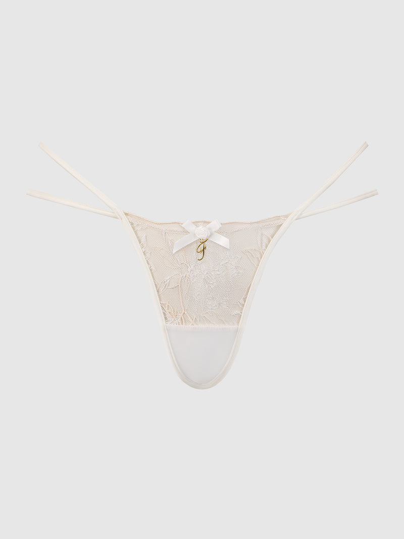 The Delfina Embroidered Backless Cheeky by Frederick's of Hollywood features a cream lace design with thin side straps, a small white bow at the center, and a gold charm beneath, shown against a plain white background.
