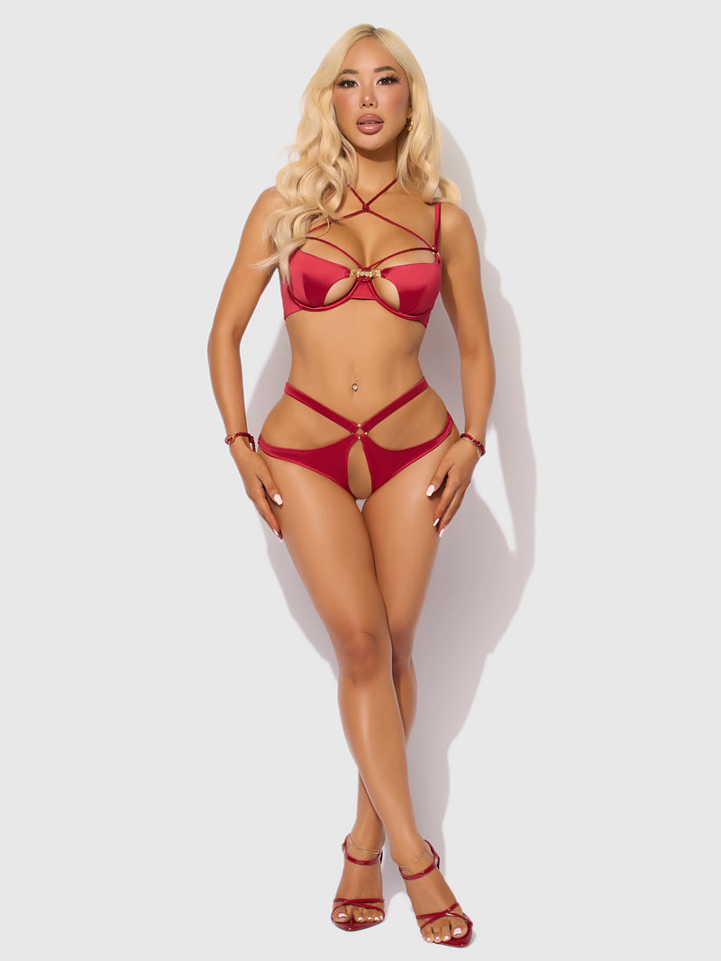 A woman with long blonde hair stands against a white background, modeling the Frederick's of Hollywood Nadya Liquid Satin & Rope Crotchless & Hand Cuff Hipster set. She faces forward, hands on her thighs, showing off its edgy red strappy details.