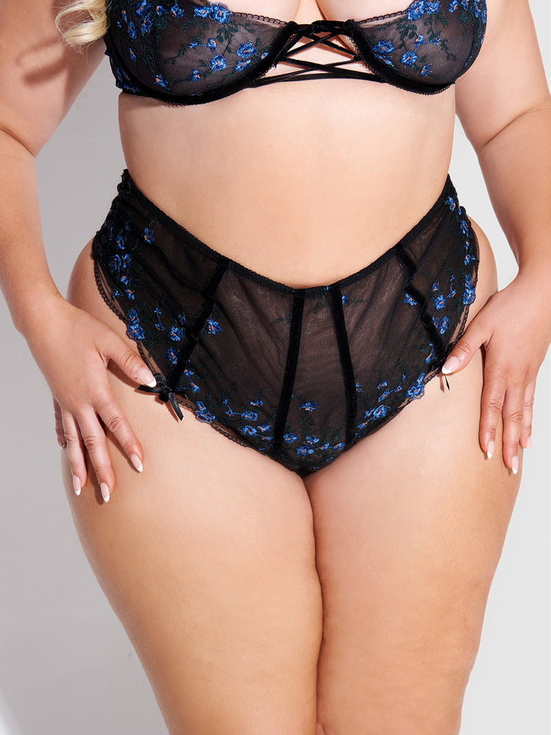 Model wears the Frederick's of Hollywood Harlow Embroidered Mesh Lace Up Backless Cheeky set—black sheer lingerie with blue floral embroidery, featuring a bra and high-waisted cheeky panties—standing hands on hips.