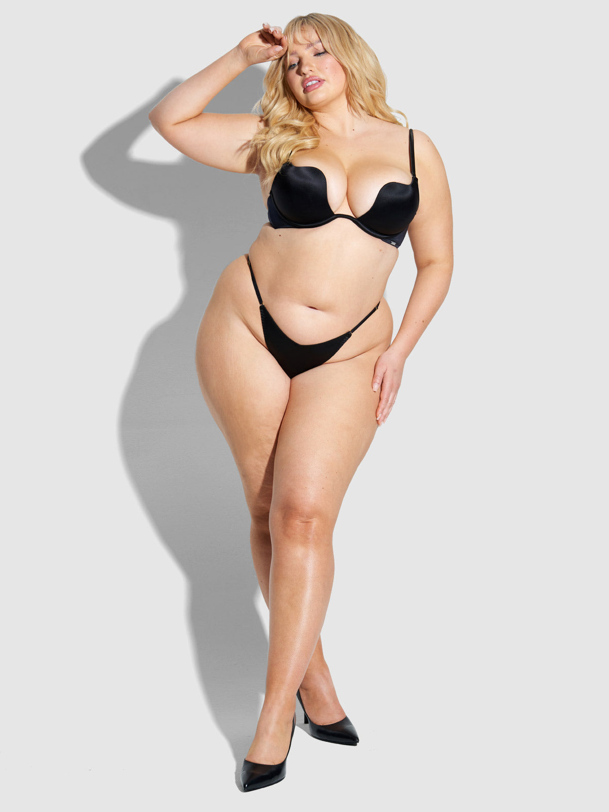 A woman with long blonde hair poses confidently in the Frederick's of Hollywood Lexi Liquid Satin G-String and high heels against a plain light gray background, one hand raised to her forehead as she gazes forward.