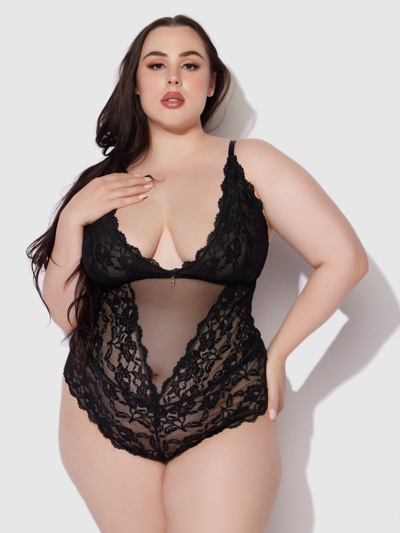 A woman poses confidently in the Mabel Wireless Teddy by Frederick's of Hollywood, a black lace piece with sheer detailing, a plunging neckline, and scalloped edges, set against a plain light background.