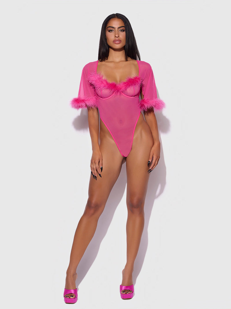 A woman with long dark hair poses confidently against a white background in the Frederick's of Hollywood Scarlett Bell Sleeved Marabou Bodysuit, a hot pink sheer mesh one-piece featuring feather trim and bell sleeves, paired with matching pink heels.