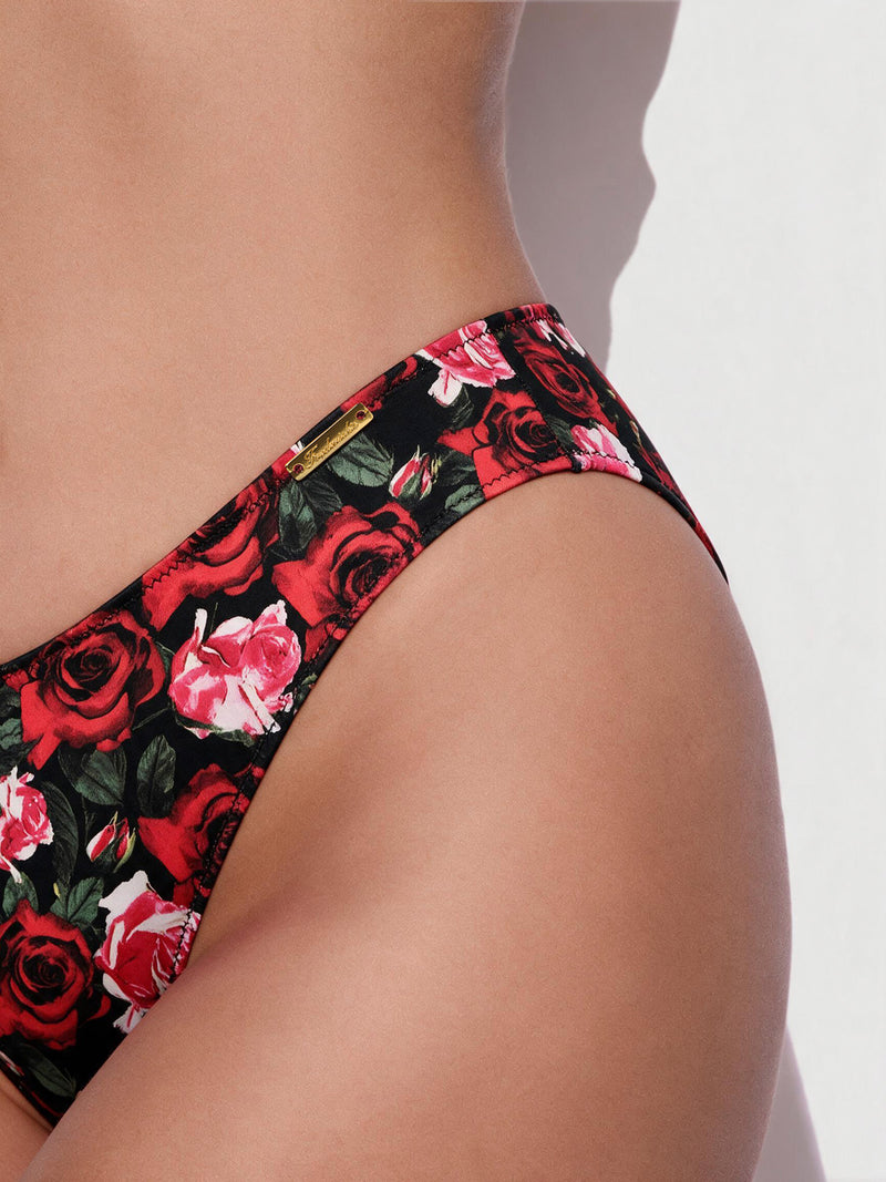 Close-up of a person’s hip and waist wearing the Frederick’s of Hollywood Lexi Liquid Satin Hipster in black, featuring a vibrant red and pink rose floral pattern against a plain light background.