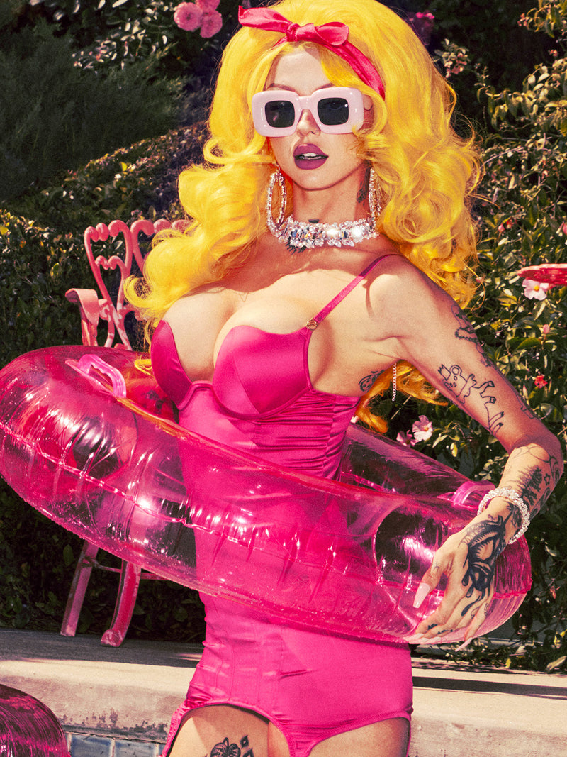 A person with bright yellow hair, in a hot pink swimsuit and bold jewelry, stands poolside with a pink inflatable ring—evoking the playful style of Frederick's of Hollywood's Rising Star Plunge Open Back Chemise Set in Petra Satin—amid pink chairs and flowers.