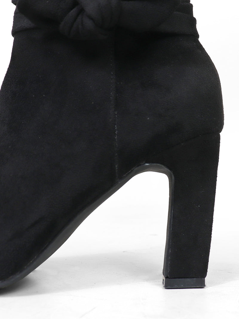 Close-up of the Janeth Ankle Heel Boot by Frederick's of Hollywood, featuring black suede, a chunky heel, and an elegant knot detail near the top. The boot is displayed against a plain white background.
