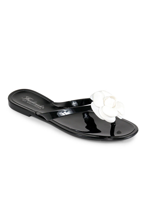 The Jelli Camellia Thong Sandals by Frederick's of Hollywood are glossy black with a white flower on the strap, featuring a flat sole and open toe‚Äîstylish, feminine footwear ideal for warm weather.