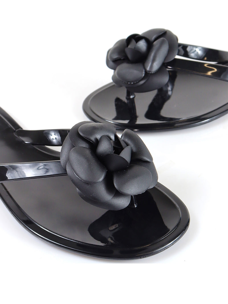 Frederick's of Hollywood Jelli Camellia Thong Sandals in shiny black feature a glossy finish and large black camellia flowers on each strap‚Äîan elegant choice for chic warm-weather looks, shown on a white background.