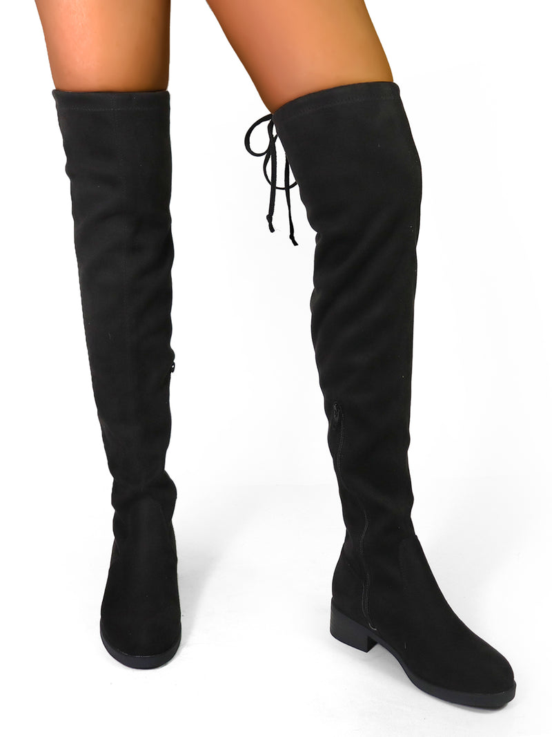 A person wearing Frederick's of Hollywood Letizia Thigh High Super Stretch Boots with low chunky heels stands on a white background. These black boots showcase a statement-making style, highlighted by the lace-up detail at the back.