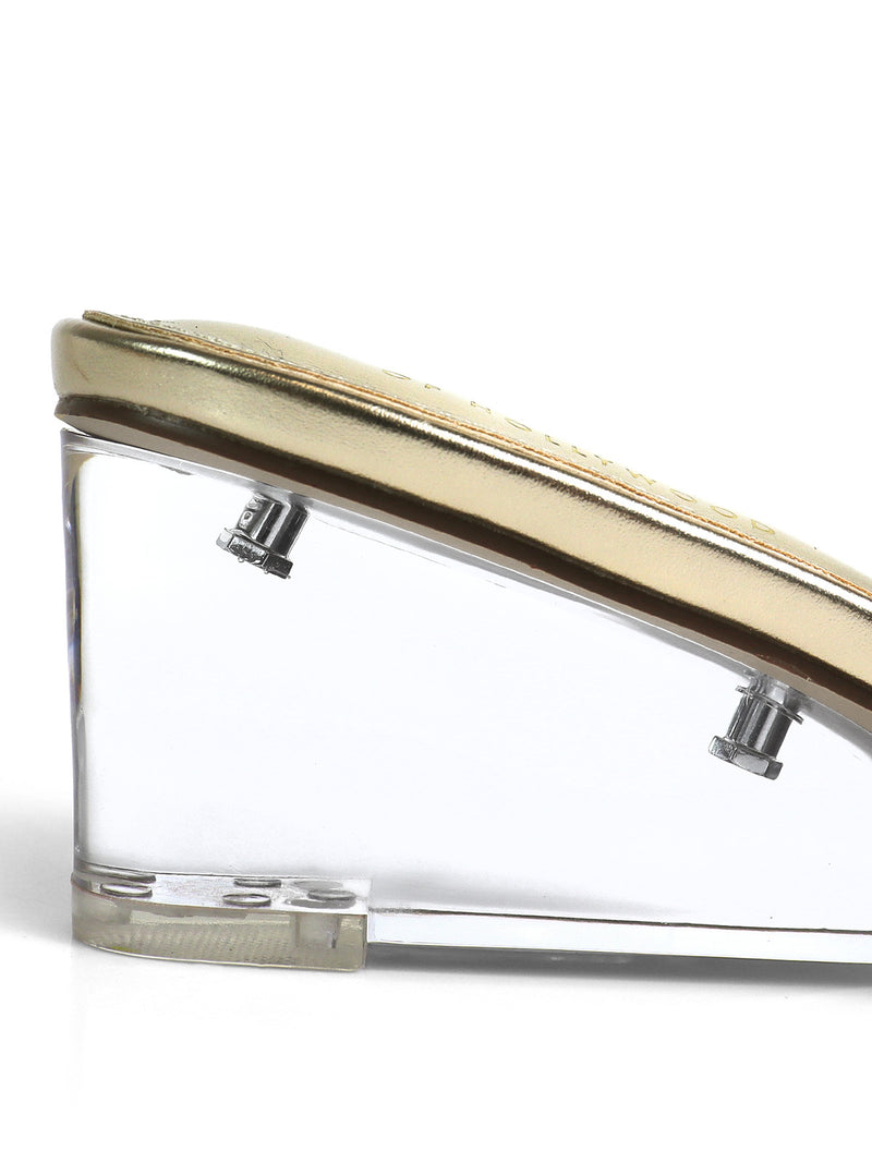 A close-up side view of the Frederick's of Hollywood Lora Transparent Mule Wedges shows a minimalist design with a metallic gold upper, a clear wedge heel featuring two visible silver screws.