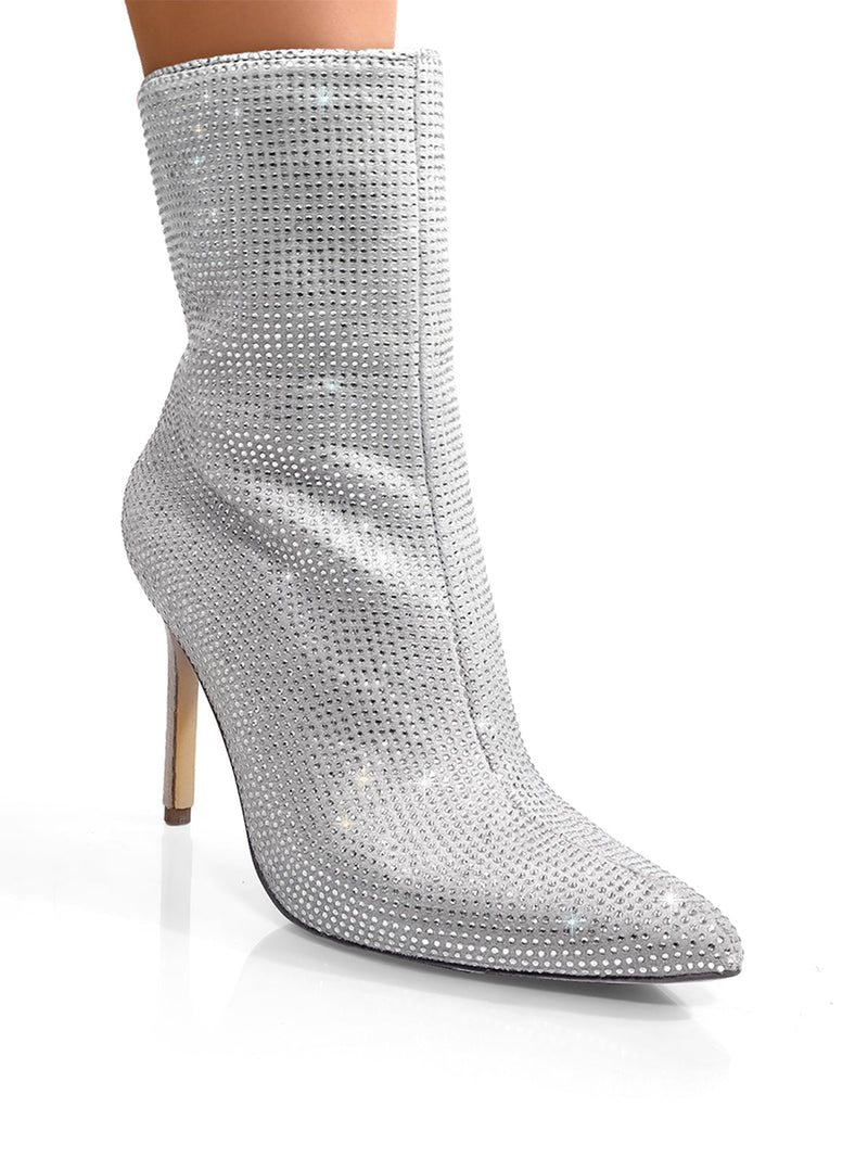 A close-up of Frederick's of Hollywood Nadia Rhinestone Booties, featuring a pointed toe and stiletto heel, sparkling on a woman's leg against a white background.