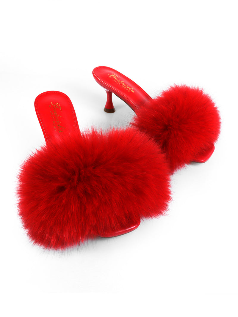 The Frederick’s of Hollywood Purr Square Toe Fur Heel is a pair of red high-heeled slippers featuring a chic square-toe silhouette and large, fluffy red pom-poms on the toes, displayed against a white background.