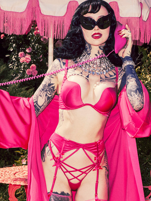 A woman wears the Frederick's of Hollywood Petra Satin Short Robe in vibrant pink, paired with bold jewelry and black sunglasses, holding a pink phone amid pink flowers and vintage-style decor.