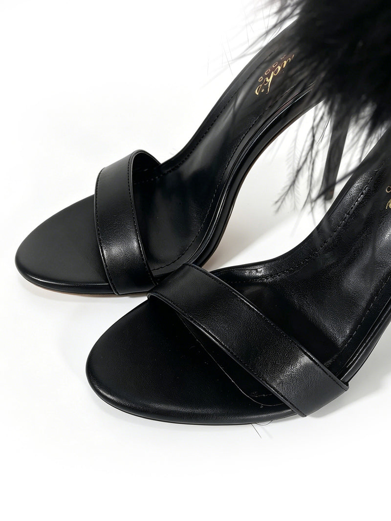 A close-up of the Raquel Feather Stiletto Heel by Frederick’s of Hollywood, featuring elegant black high-heeled sandals with a single toe strap and fluffy feather detail near the ankle, showcasing a sleek stiletto silhouette that captures the essence of modern luxury, set against a white background.