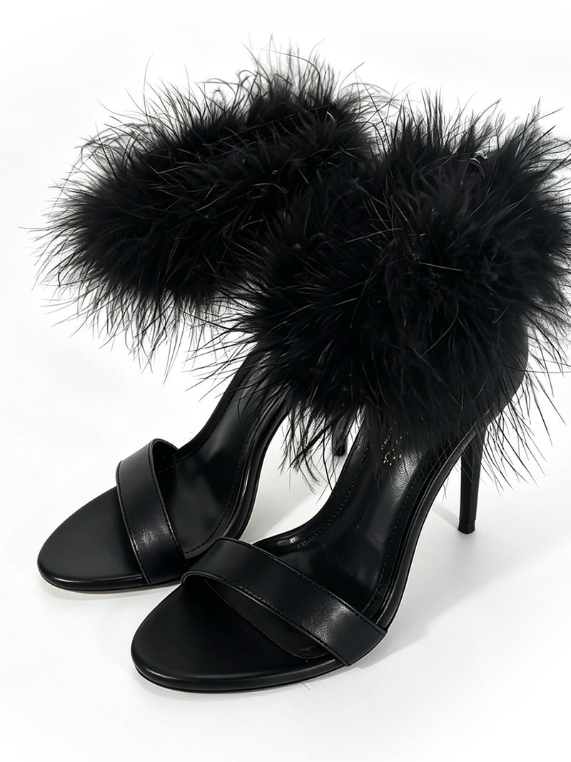 The Raquel Feather Stiletto Heel by Frederick’s of Hollywood features a striking black stiletto silhouette with minimalist front straps and bold, fluffy black feather accents around the ankle straps, embodying modern luxury as they are displayed against a white background.