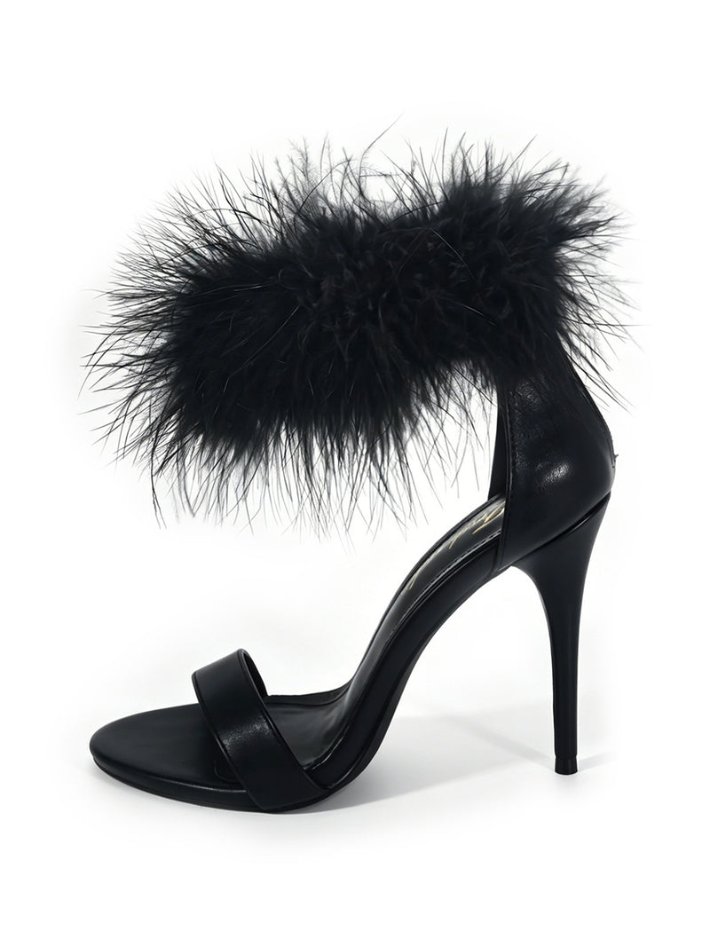 The Raquel Feather Stiletto Heel by Frederick’s of Hollywood features a sleek black design with an ankle strap accented by plush black feathers, a single toe strap, and a slender stiletto heel. Showcased on a white background, this elegant shoe embodies modern luxury with its striking silhouette.