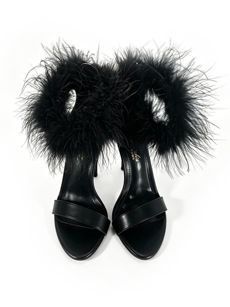 Introducing the Raquel Feather Stiletto Heel by Frederick’s of Hollywood: a pair of black high-heeled sandals with an elegant stiletto silhouette, adorned with fluffy black feather ankle straps and sleek toe straps, showcased on a pristine white background—a bold statement of contemporary luxury.