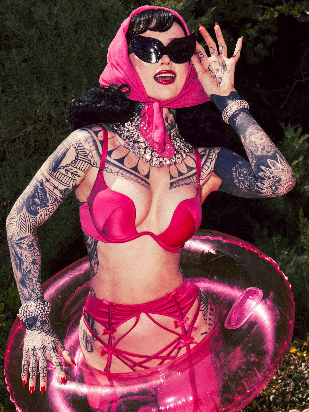 A tattooed model in the Frederick's of Hollywood Rising Star Plunge Push Up Bra in Petra Satin, with convertible straps, poses outdoors in vintage pin-up style, accessorized with a garter belt, headscarf, cat-eye sunglasses, and pink inflatable ring.