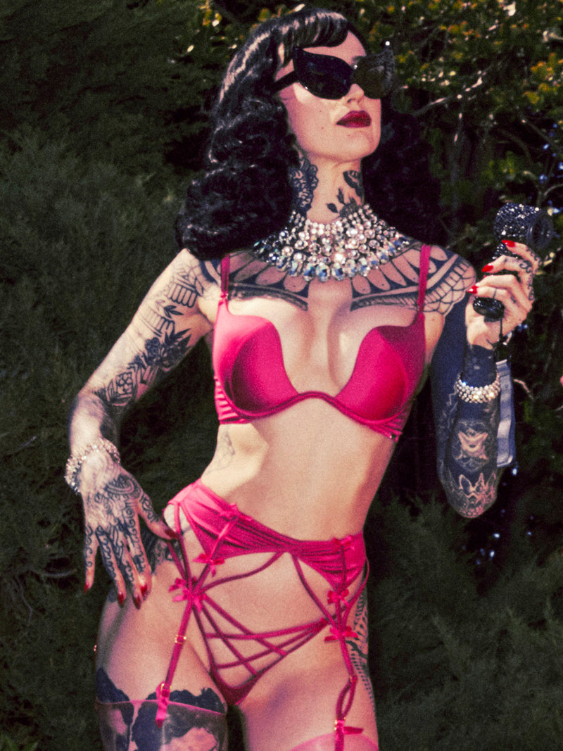 A tattooed person with wavy black hair and sunglasses poses outdoors, holding a small microphone. They wear a red Frederick’s of Hollywood Petra Satin & Mesh Garter Belt and a jeweled necklace, with green foliage in the background.