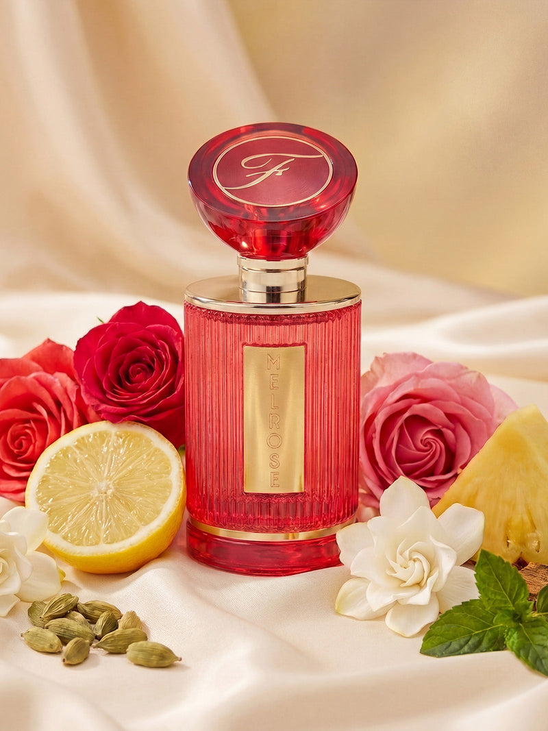 A pink Melrose 60ml Eau De Parfum by Frederick's of Hollywood rests on satin, surrounded by lemon, pineapple, cardamom pods, roses, white flowers, and mint—evoking a vibrant floral scent with amber musk notes.