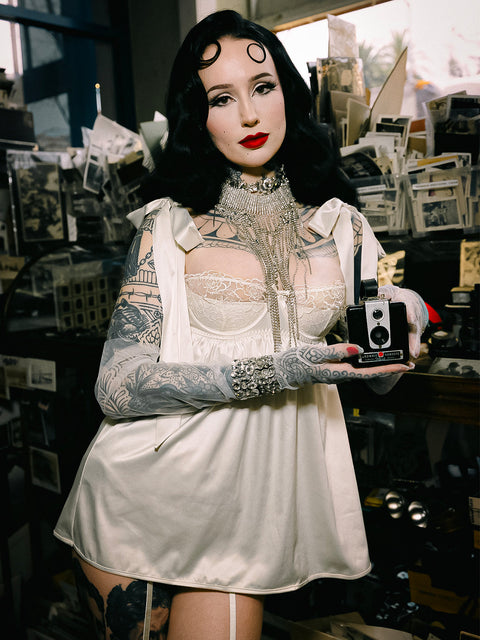 A dark-haired, tattooed woman with red lipstick wears the Frederick's of Hollywood Melrose Satin & Embroidered Lace Underwire Babydoll Set and ornate jewelry, standing in a cluttered vintage room holding a small box camera.