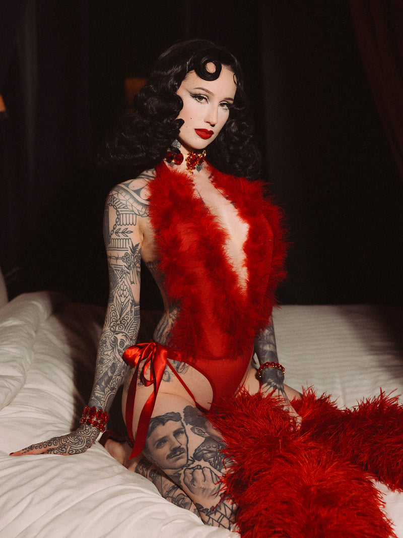 A person with vintage black hair, bold makeup, and tattoos poses on a bed in glamorous boudoir lighting, wearing the Scarlett Plunging Drop Off Marabou Teddy by Frederick's of Hollywood.