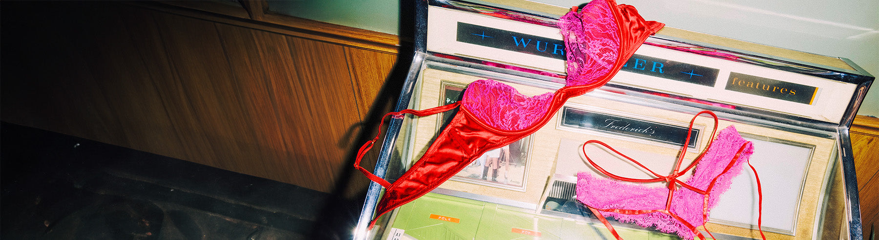 A pink satin and lace lingerie set displayed on a vintage glass-front cabinet against a dark wood-paneled wall.