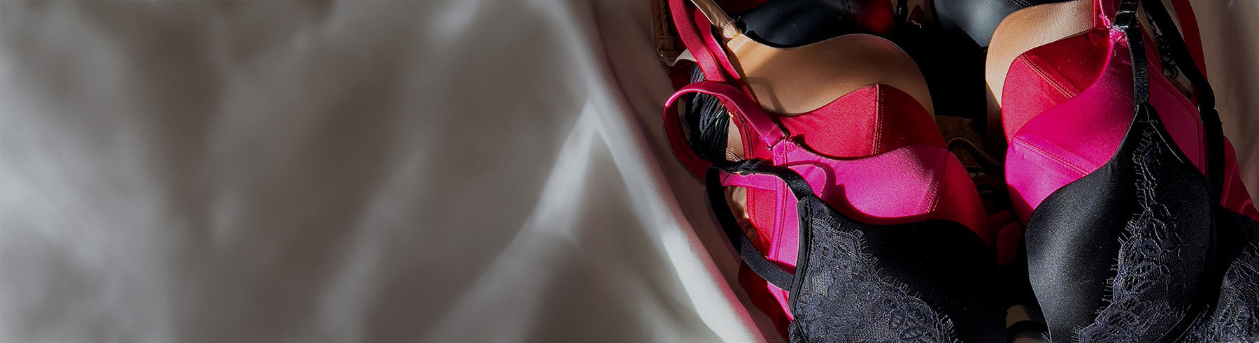 A pile of women's lingerie bras in bright pink and black, with one black lace bra partially visible on the left, set against a soft white fabric background.