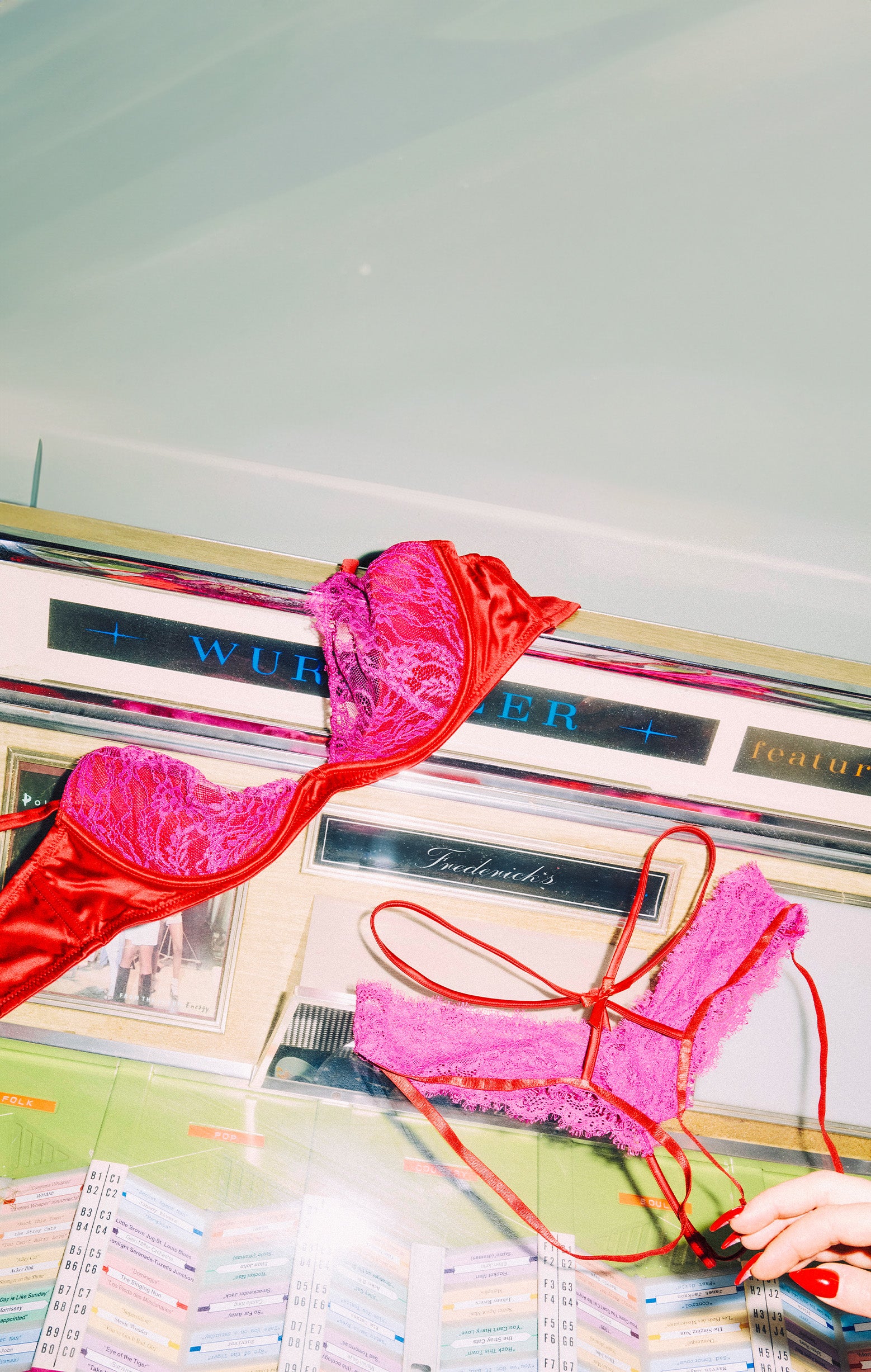 A vintage-style lingerie display or counter scene with two bras draped over a counter: one bright red satin bra and one hot pink lace bra, with a hand holding a phone in the lower right corner.