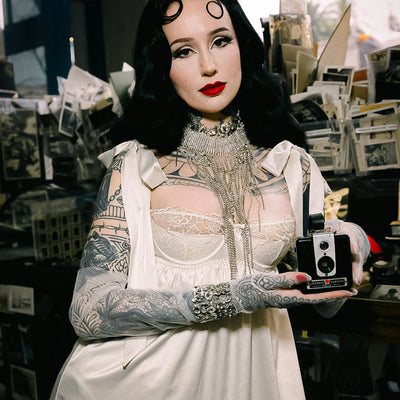A tattooed woman in a white satin dress and gloves holds a vintage camera. She has dark hair styled in waves, wears bold red lipstick, and stands in front of a cluttered background with scattered photos and papers.