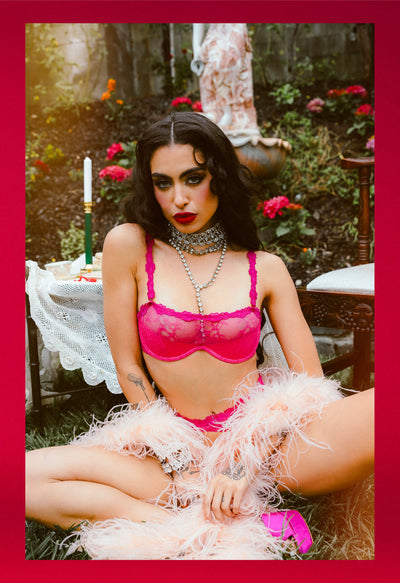 Woman in a pink bra and feathered boa sitting outdoors with flowers and furniture in the background.