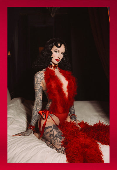Person in a red outfit with feathered details on a bed, against a dark background.