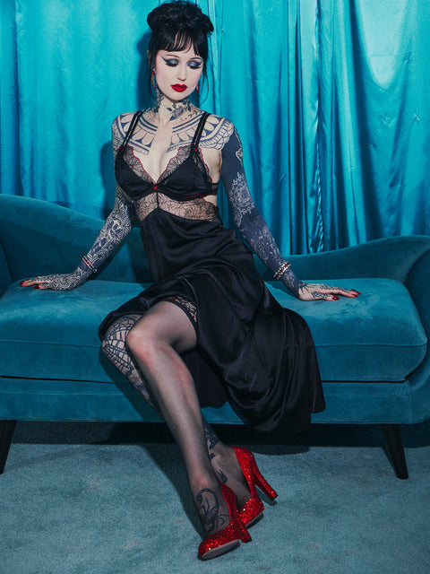 A woman with dark hair and tattoos sits on a blue couch, wearing the Ilaria Embroidered Tulle Triangle Top Long Gown by Frederick's of Hollywood and red glitter high heels, in front of turquoise curtains.