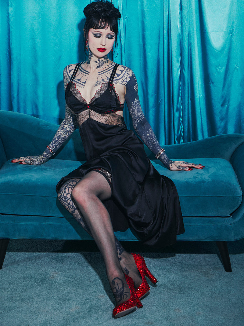 A woman with dark hair and tattoos sits on a blue couch, wearing the Ilaria Embroidered Tulle Triangle Top Long Gown by Frederick's of Hollywood and red glitter high heels, in front of turquoise curtains.